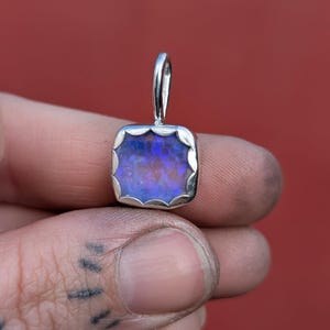 May include: A square pendant featuring a vibrant blue and purple iridescent opal stone set in a polished silver frame with a scalloped edge and a silver bail at the top for attachment to a chain or cord.