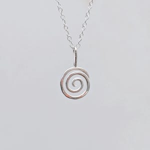 May include: A silver pendant necklace with a spiral design. The pendant, a small open spiral, hangs from a fine chain. The necklace is shown against a plain, light background. The chain and pendant appear to be made of silver.