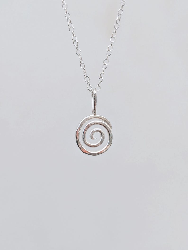 May include: A silver-colored pendant necklace featuring a spiral design. The pendant is a small, open spiral shape suspended from a delicate chain. The necklace is displayed against a plain, light background.