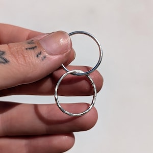 Może przedstawiać: Dwa srebrne pierścionki, jeden gładki i jeden teksturowany, są prezentowane na białym tle. Pierścionki są okrągłe i wydają się być wykonane z metalu. Teksturowany pierścionek ma fasetowaną powierzchnię.