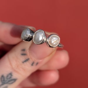 May include: A silver ring featuring three irregularly shaped, off-white pearls. The pearls are set in individual silver bezels, creating a unique and organic design. The ring is shown against a red background.