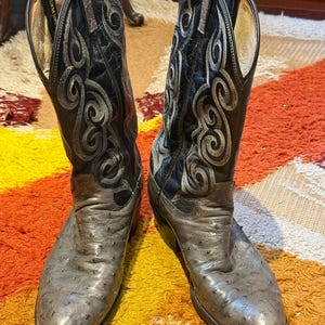 May include: A pair of cowboy boots with a distressed, textured gray leather foot and a black leather upper. The upper features intricate silver embroidery. The boots are displayed on a colorful, patterned rug.