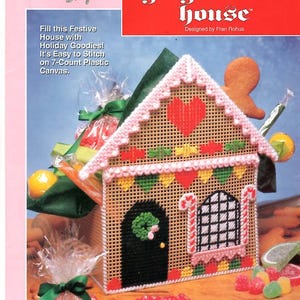 May include: A plastic canvas gingerbread house craft kit package featuring a tan house with a black door, pink roof trim, a red heart, and candy cane window frames. The text reads Plastic Canvas, The Needlecraft Shop, and gingerbread house.
