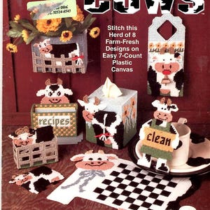 May include: A plastic canvas craft pattern booklet titled Country Cows by The Needlecraft Shop. The cover displays eight cow-themed projects including a wall pocket, tissue box cover, recipe holder, and coasters, all stitched with black and white yarn.