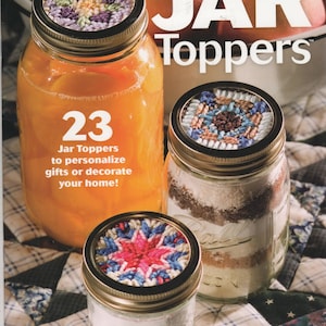 May include: A craft book cover titled Plastic Canvas JAR Toppers featuring three glass jars with embroidered circular lids. The text reads 23 Jar Toppers to personalize gifts or decorate your home! The Needlecraft Shop logo is in the corner.