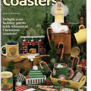 May include: A craft pattern book titled Plastic Canvas Christmas Coasters featuring designs by Vicki Blizzard. The cover displays various plastic canvas projects including a praying angel, a fireplace with logs, a festive hat, and a snack tray.