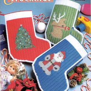 May include: A craft pattern book titled Plastic Canvas Christmas Stockings by Annie's Attic, featuring three handmade stockings: a red one with a Christmas tree, a green one with a reindeer, and a blue one with a Santa Claus figure.