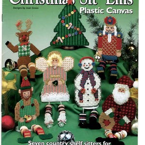 May include: A craft pattern book titled Country Christmas Sit 'Ems Plastic Canvas featuring seven plastic canvas figures including a reindeer, Christmas tree, nutcracker, angel, snowman, elf, and Santa Claus with button accents and yarn details.