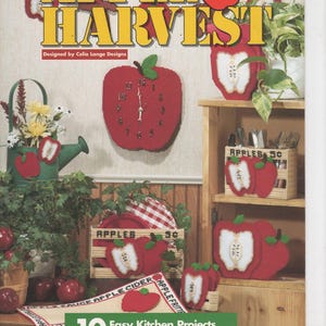 May include: A pattern book titled Apple Harvest featuring plastic canvas projects. The cover displays red apple-shaped clocks, containers, and coasters with white apple slice details. Text includes 10 Easy Kitchen Projects You'll Love to Stitch.