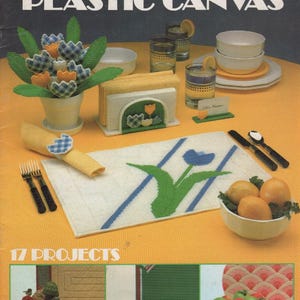 May include: A Leisure Arts craft leaflet titled Table Settings for Plastic Canvas, featuring 17 projects. The cover displays a table with a plastic canvas tulip bouquet, napkin holder, place mat, and napkin ring, alongside bowls of fruit.