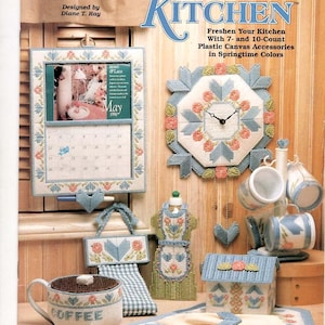 May include: A plastic canvas craft pattern book titled The Needlecraft Shop Hearts & Flowers Kitchen featuring projects like a calendar, wall clock, mug tree, coffee mug, apron-shaped wall hanging, and a small house-shaped box in pastel colors.