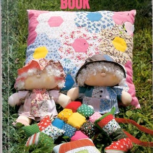 May include: A book cover with the title "Patchwork & Quilting Book" in pink letters. Two handmade dolls and a patchwork pillow are in the foreground. A collection of small, colorful patchwork items are scattered in front of the dolls, all on a bed of green grass.