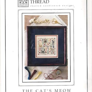 May include: A framed cross-stitch sampler with a blue border, featuring an alphabet and cat motifs. The text "THE DRAWN THREAD" and "THE CAT'S MEOW" are visible. Additional craft supplies like scissors and thread are included.