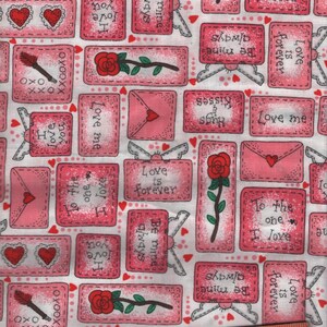 May include: A white fabric featuring a repeating pattern of pink rectangular motifs containing red roses, envelopes, and text including "Love is forever," "Be mine always," and "To the one I love." A wooden ruler shows a 6-inch measurement.