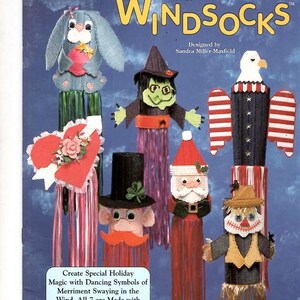 May include: A craft pattern booklet titled The Needlecraft Shop Plastic Canvas Windsocks featuring seven designs including a bunny, a heart, a witch, a leprechaun, Santa Claus, an eagle, and a scarecrow, all created with 7-count plastic canvas.