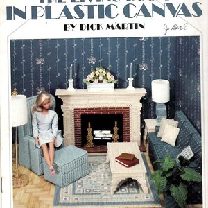 May include: A Leisure Arts craft leaflet titled The Living Room in Plastic Canvas by Dick Martin, featuring a miniature dollhouse living room scene with a fireplace, furniture, a doll, and accessories constructed from plastic canvas.