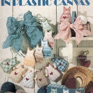 May include: A craft instruction leaflet titled Leisure Arts Leaflet 1185 Homespun Cats in Plastic Canvas. The cover displays various cat-shaped plastic canvas projects in pastel colors with heart motifs, alongside yarn balls, a wreath, and a basket.