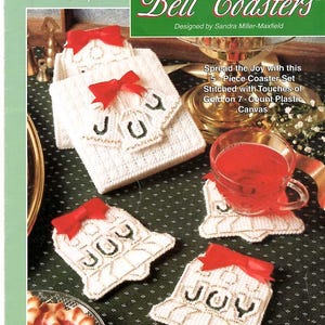 May include: A plastic canvas craft kit for Bell Coasters featuring white bell-shaped coasters with gold trim, green stitched lettering spelling JOY, and red ribbon bows. The packaging includes the text The Needlecraft Shop and Bell Coasters.