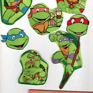May include: A collection of seven Teenage Mutant Ninja Turtles paper cutouts featuring various characters in green, blue, orange, red, and purple, positioned above a wooden ruler measuring 5 inches in length with Keebler and American Greetings text.
