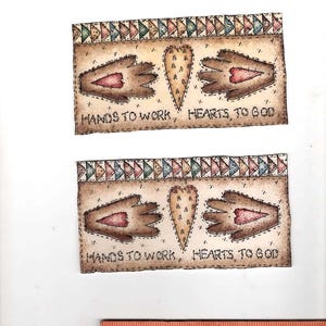 May include: Two rectangular paper cutouts featuring a tan background with a border of colorful triangles. Each cutout displays two brown hand shapes, a central heart, and the text "HANDS TO WORK, HEARTS TO GOD". A wooden ruler shows a width of 5 inches.