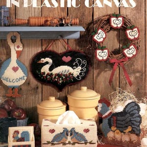 May include: A Leisure Arts craft leaflet titled More Country Things in Plastic Canvas, featuring various plastic canvas projects including a goose welcome sign, a heart-shaped swan wall hanging, an apple wreath, and a tissue box cover.