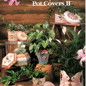 May include: A craft pattern book titled Annie's Attic Plastic Canvas Pretty Plant Pot Covers II featuring various decorative plastic canvas covers for potted plants, including designs with butterflies, rabbits, and floral motifs in pastel colors.
