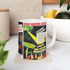 May include: White ceramic mug featuring a vintage movie poster design with the text "FLYING SAUCERS ATTACK!" and "EARTH vs. THE FLYING SAUCERS." The design includes images of flying saucers, people, and the words "WARNING! TAKE COVER!"