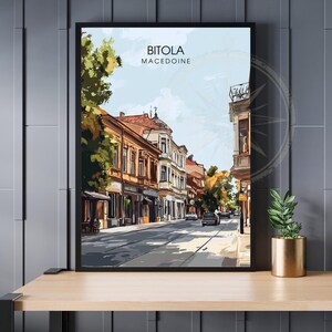 Peut inclure: Une impression d'art encadrée représentant une scène de rue à Bitola, Macédoine. L'œuvre présente des bâtiments aux façades orange et blanches, une rue bordée d'arbres et un ciel bleu. Le texte "BITOLA MACEDOINE" est en haut.