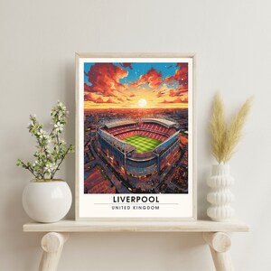 May include: A framed print of an aerial view of Anfield Stadium in Liverpool, England. The stadium is surrounded by a red brick wall and a green field. The sky is a vibrant orange and red, with a bright sun shining in the distance. The text "LIVERPOOL" and "UNITED KINGDOM" is printed below the image.