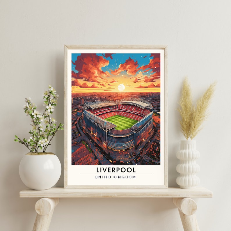May include: A framed print of an aerial view of Anfield Stadium in Liverpool, England. The stadium is surrounded by a red brick wall and a green field. The sky is a vibrant orange and red, with a bright sun shining in the distance. The text "LIVERPOOL" and "UNITED KINGDOM" is printed below the image.