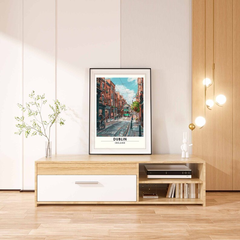 May include: A framed print depicting a street in Dublin, Ireland, with a green tram and red brick buildings. The print is on a light wood cabinet with a white drawer. A vase with greenery and a decorative light fixture are also visible.