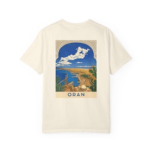 Peut inclure: Un t-shirt à manches courtes de couleur crème avec un motif rectangulaire au dos. L'illustration représente une vue côtière de la ville d'Oran encadrée par une arche, montrant un port bleu, des bâtiments et un cactus, avec le texte ORAN en bas.