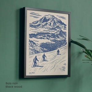 May include: A framed print depicting a mountain scene with skiers, rendered in blue and white. The print is set within a black wooden frame. The text "Les Alpes" is visible. The frame is mounted on a green wall.