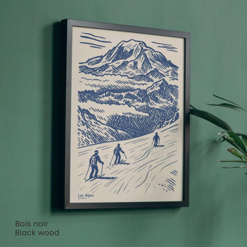 May include: A framed print depicting a mountain scene with skiers, rendered in blue and white. The print is set within a black wooden frame. The text "Les Alpes" is visible. The frame is mounted on a green wall.