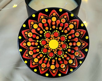 May include: A black candle tin with a lid featuring a hand-painted mandala design in vibrant shades of yellow, orange, and red. The circular lid displays a symmetrical pattern of dots and curved shapes. The candle inside is cream-colored.