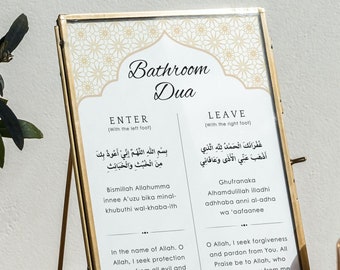 May include: A gold-framed sign with the words "Bathroom Dua" in a decorative font. The sign has two columns, one labeled "Enter" and the other labeled "Leave". Each column contains Arabic text and an English translation. The sign is on a table with a vase of greenery and a candle.