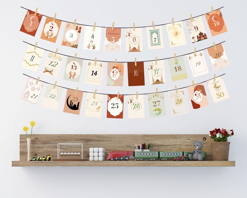 May include: A string of 30 cards with gold numbers and Islamic-themed illustrations, hanging on a white wall. The cards are decorated with crescent moons, stars, lanterns, and mosques. The cards are numbered 1 through 30, with the word "Ramadan" on the fifth card.