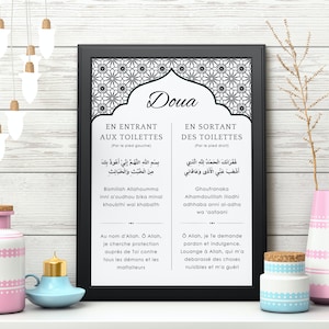 Puede incluir: Una impresión enmarcada con texto árabe, con un marco negro y un fondo blanco. La impresión está decorada con un diseño floral en la parte superior. Objetos decorativos, incluyendo una botella rosa y blanca, un candelabro azul y un jarrón rosa, están en primer plano.