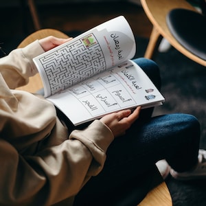 Może przedstawiać: Osoba siedzi na krześle i czyta książkę z labiryntem i grą słowną. Książka nosi tytuł "Gra Ramadanowa", a gra słowna nosi tytuł "Gra słowna". Książka jest w języku arabskim.
