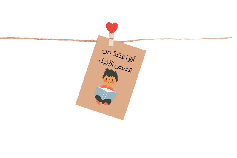 May include: A brown card with a red heart clothespin hanging from a string. The card has a cartoon illustration of a boy reading a book and the text "Aقرأ قصة من فصص الأنبياء" in Arabic.