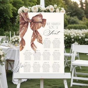 May include: A wedding seating chart sign on a white easel featuring a large watercolor brown ribbon illustration and the text "WE SAVED YOU A SEAT" followed by a list of ten tables with names printed in black ink on a cream-colored background.