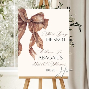 Puede incluir: Un letrero de bienvenida para una despedida de soltera, con una ilustración de un lazo marrón. El letrero dice "She's Tying The Knot" y "Welcome to ABAGAIL'S Bridal Shower" con la fecha 11.03.26.