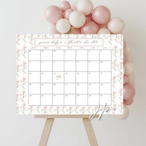 May include: A white calendar with a floral design and the text "guess baby's Smith's due date." The calendar is displayed on a wooden easel, with pink and white balloons in the background. The calendar has a grid of dates.