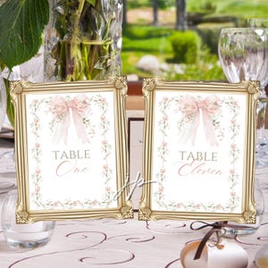 May include: Two framed table number signs with gold ornate frames. Each sign features a floral design with a pink bow and the words "TABLE One" and "TABLE Eleven" in elegant script. The signs are set on a table with a white tablecloth.
