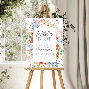 May include: A rectangular sign featuring a white arched center with the text Wildly In Love, Welcome To Samantha's Bridal Shower. The border is decorated with a colorful watercolor illustration of various wildflowers, greenery, and small blossoms.