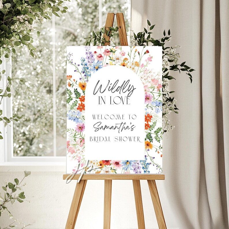 May include: A rectangular sign featuring a white arched center with the text Wildly In Love, Welcome To Samantha's Bridal Shower. The border is decorated with a colorful watercolor illustration of various wildflowers, greenery, and small blossoms.