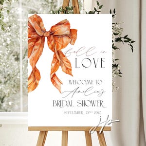 Pode incluir: Uma placa branca em um cavalete de madeira com o texto "Fall in Love" e "Welcome to Amelia's Bridal Shower September 22nd 2025". A placa apresenta uma ilustração de um laço laranja.