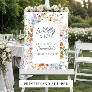 May include: A rectangular sign on a white easel featuring a colorful watercolor floral border with orange, pink, blue, and white flowers. The text reads Wildly IN LOVE WELCOME TO Samantha's BRIDAL SHOWER in a mix of script and sans-serif fonts.