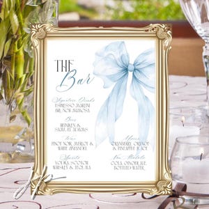 May include: A gold framed sign with the text "The Bar" and a light blue bow illustration. The sign lists signature drinks, beers, wines, spirits, mixers, and non-alcoholic beverages. The sign is on a table with candles.