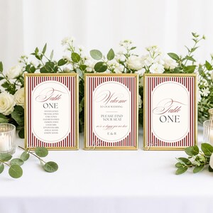 Puede incluir: Tres letreros con marco dorado y fondo de rayas verticales en rojo oscuro y blanco. Los letreros presentan centros ovalados blancos con texto en negro y rojo, incluyendo Table ONE, Welcome to our wedding y una lista de nombres.